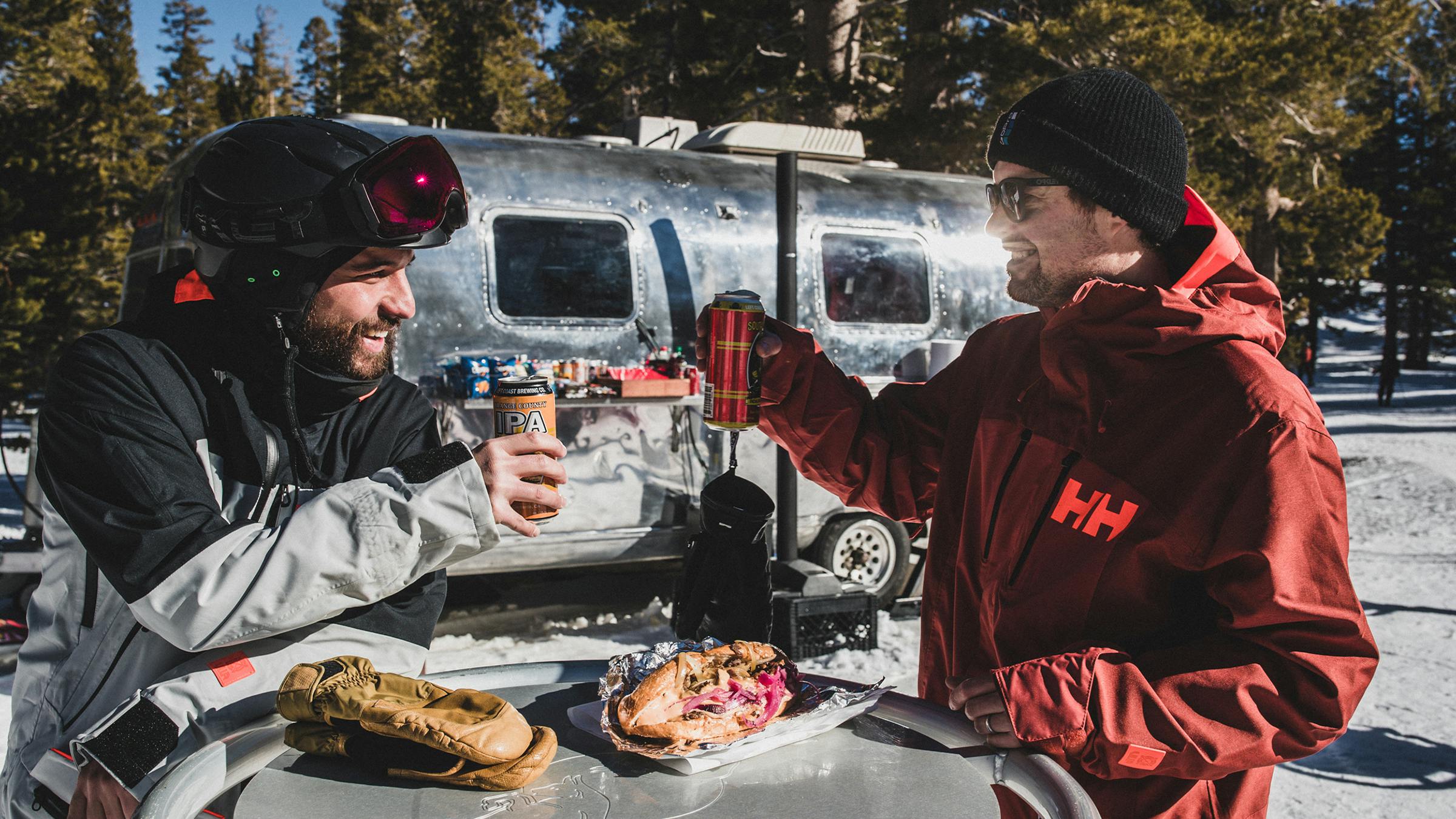 Two men in ski gear holding drinks with cheesesteaks on the table