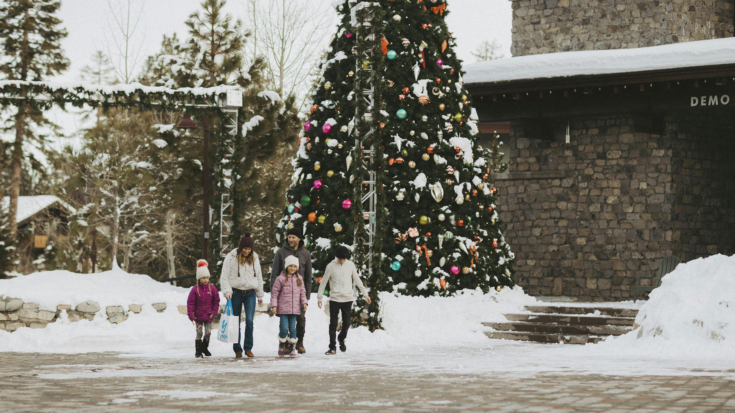 Family walking through snowy Village at Mammoth with decorated Christmas tree behind them