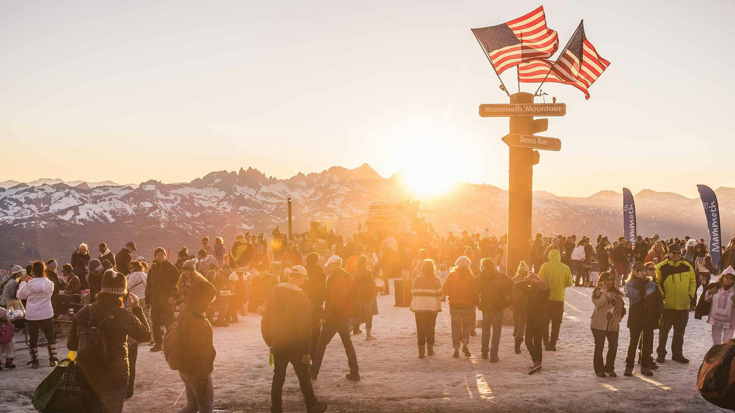 View of sun setting over Minarets from the top of Mammoth Mountain with American flags on top of the summit sign and crowd of people watching the sun set