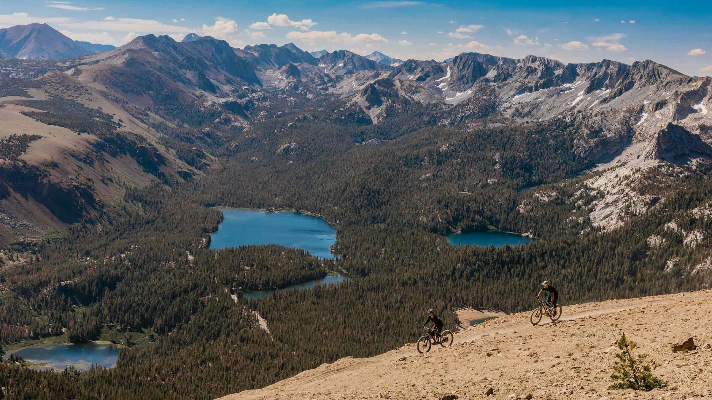 Two mountain bikers riding Off the Top with a view of the Lakes Basin.