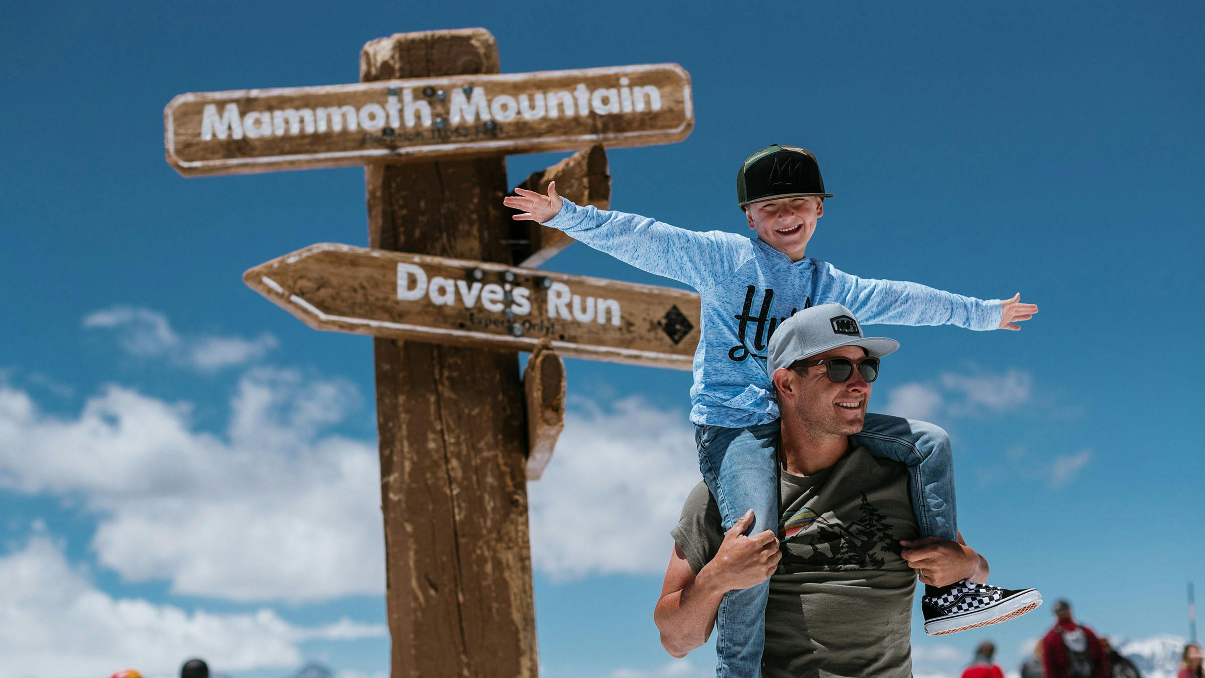 Boy on father's shoulders with outstretched arms in front of Mammoth Mountain summit sign